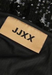 Tessuto nero con paillettes e un'etichetta beige che mostra "JJXX" in lettere nere, cucita lungo il bordo del tessuto.