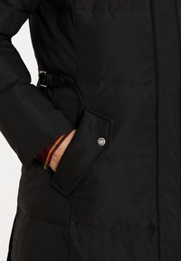 Melnā polsterētā jaka ar sānu kabatu un spiedpogām. Tam ir jostas viduklis un redzama sarkana un dzeltena šķautņu oderes līnija pie piedurknēm.