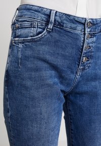 Los jeans de mezclilla azul oscuro presentan un cierre de cinco botones, un diseño de bolsillo tradicional y un ligero efecto de desgaste, mostrando un estilo casual clásico.