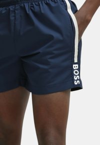 Pantaloni da bagno blu navy realizzati in tessuto leggero, con strisce bianche sui lati e un logo stampato sulla parte bassa della gamba. Include una cintura elastica.