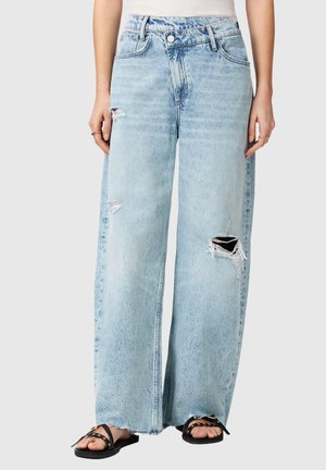 Hellblaue Jeans mit weitem Bein und ausgefransten Rissen an den Beinen, kombiniert mit schwarzen offenen Sandalen und einem weißen Oberteil, gezeigt von der Taille bis zu den Füßen.