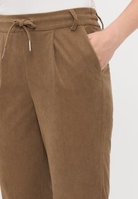 Brune fløjlbukser med snor i taljen, plisseret front og sidelomme, med en ribbet tekstur. Nærbillede af taljebåndet.