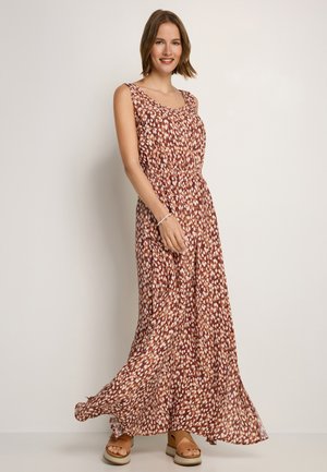 Lächelnde Frau, die ein ärmelloses, braun-weiß gemustertes Maxikleid und hellbraune Plateausandalen trägt, steht vor einer schlichten hellen Wand.