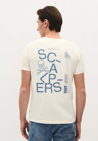 Korte mouwen crème t-shirt met een blauwe grafische print met tekst, schedel en gekruiste botten op de achterkant. Zacht katoenen materiaal.