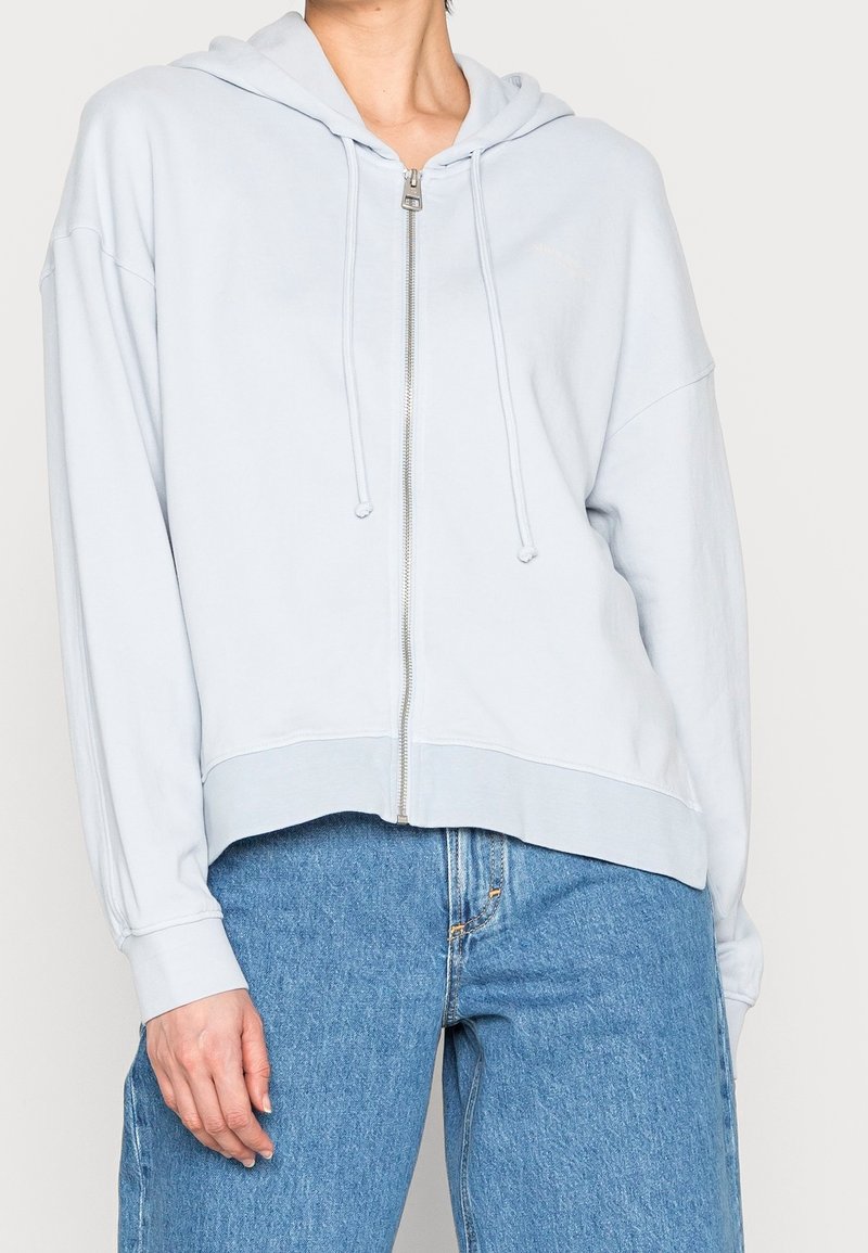 Sudadera azul claro con cremallera, de textura suave, corte relajado y capucha con cordón, combinada con jeans de mezclilla de talle alto.