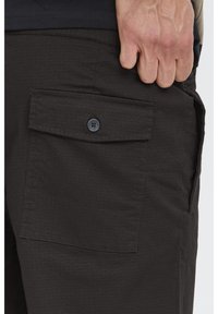 Pantalones cortos cargo de color gris oscuro con tela texturizada, con un bolsillo con solapa asegurado con un botón en el lado derecho y una mano visible dentro del bolsillo.