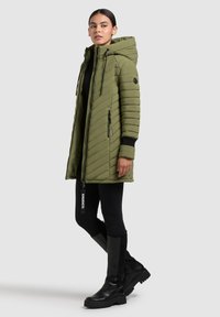 Parka verde imbottito con cappuccio, dotato di tasche con zip e rifiniture nere, abbinato a leggings neri e stivali sopra il ginocchio.