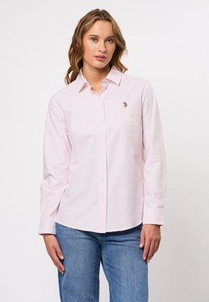 Vrouw met golvend haar die een lichtroze blouse met knopen draagt en een klein geborduurd logo, gecombineerd met een blauwe spijkerbroek, staande tegen een lichte achtergrond.