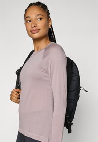 Hellbluse mit langen Ärmeln in lila mit einem strukturierten Muster und einem runden Ausschnitt, kombiniert mit einem schwarzen gepolsterten Rucksack.