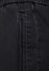 Czarny materiał dżinsowy z elastycznym pasem i pionową boczną kieszenią ozdobioną szczegółowym przeszyciem.