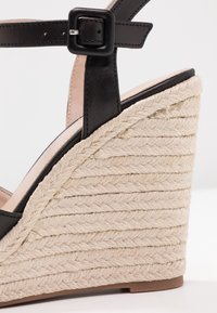 Sandale compensée avec bride de cheville en cuir noir et talon en corde tressée beige, avec une boucle carrée noire.