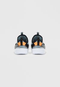 Un paio di sneakers grigie senza lacci con tiranti neri e arancioni, su suole bianche, viste da dietro su uno sfondo bianco semplice.