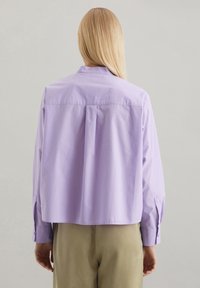 Camicia corta di un viola chiaro con colletto a camicia, maniche lunghe e una piega centrale sul retro. Tessuto liscio, fit rilassato.