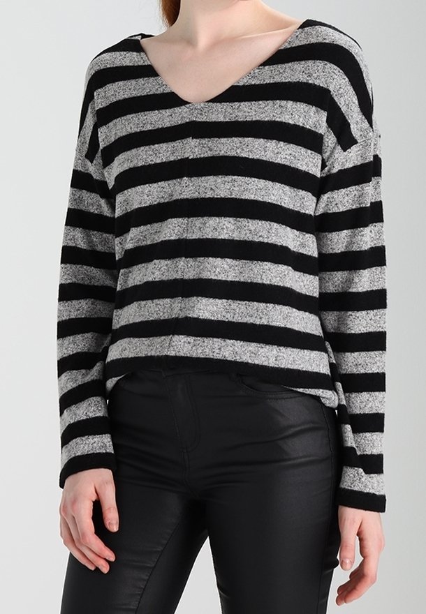 Femme portant un pull à rayures horizontales grises et noires avec un col en V et un pantalon noir ajusté, debout devant un fond uni.