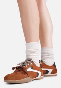 Brune semskede og lædersneakers med en hvid accent. Har flade snørebånd og en gummisål; parret med ribbede hvide sokker.