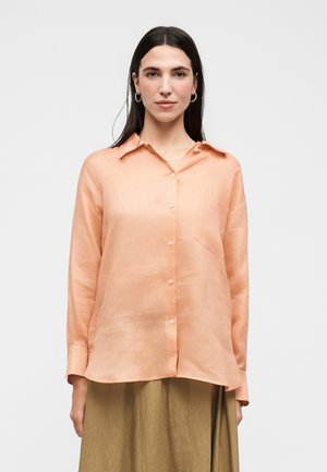 Žena s dlhými tmavými vlasmi má na sebe broskyňovú košeľu na gombíky a olivovo zelenú sukňu, stojí na jednoduchom svetlom pozadí.