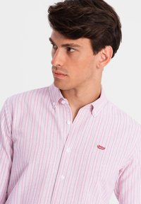 Knoopshirt in lichtroze met witte en blauwe verticale strepen. Heeft een kraag en een klein rood logo op de borst. Van katoen.