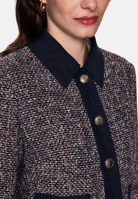 Woven jacket in navy, red, and cream with a collared neckline, gold buttons, and a textured fabric. Features a front pocket.