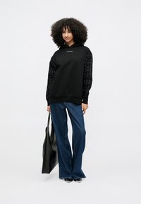 Νεαρή γυναίκα με σγουρά μαλλιά, φορώντας μαύρο φούτερ με εμπριμέ μανίκια, τζιν παντελόνι φαρδύ στα πόδια, μαύρα μυτερά παπούτσια και κρατώντας μια μαύρη τσάντα tote.