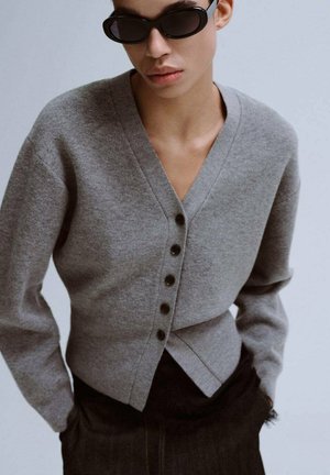Cardigan gris tricoté avec col en V, boutons sur le devant et manches longues, associé à un denim foncé. Présente un design portefeuille et une texture douce.