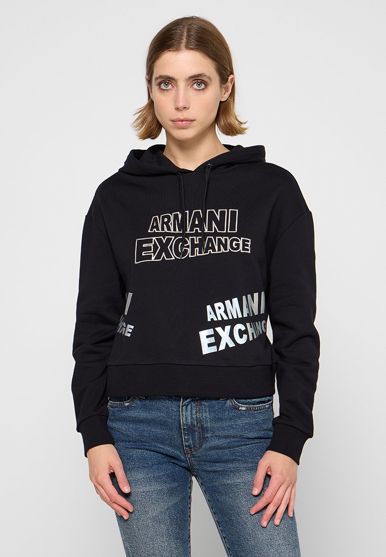 Mujer joven con cabello corto, vistiendo una sudadera negra de Armani Exchange y jeans azules, de pie contra un fondo claro y liso.