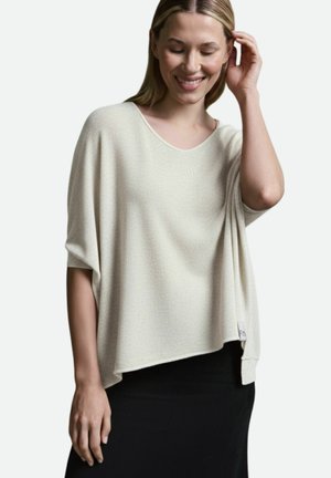 Frau lächelnd mit der Hand im Haar, trägt ein lockeres cremefarbenes Kurzarmoberteil und einen schwarzen Rock vor einem einfarbigen Hintergrund.