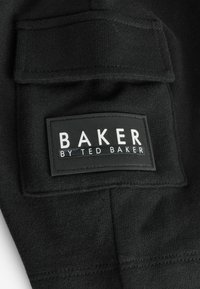 Čierna látková nášivka s nápisom "BAKER BY TED BAKER" v bielej farbe. Má obdĺžnikový tvar a textúrovaný povrch, pripevnená na odev.