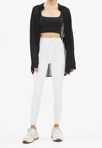 Vita högmidjade leggings med snörning framtill, svart crop top och genomskinlig svart skjorta med knappar; matchat med vita sneakers.