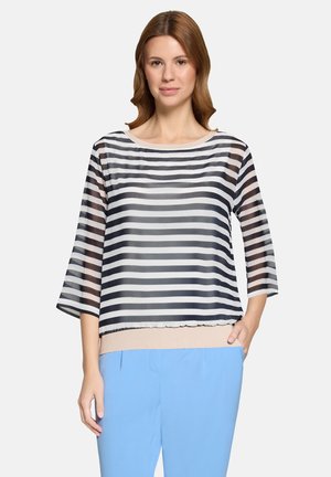 Woman wearing a black and white horizontal striped sheer blouse with beige trim and light blue pants, standing with one hand in pocket.