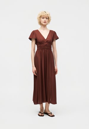 Žena s krátkymi kučeravými blond vlasmi, oblečená v stredne dlhej šaty s výstrihom do V v hrdzavej farbe a čiernych plochých sandáloch, stojaca na jednoliatej pozadí.