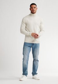 Suéter de cuello alto beige claro, silueta ajustada, textura suave. Combinado con jeans azul claro y zapatillas blancas, de pie contra un fondo llanos.