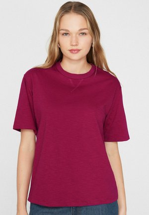 Mladá žena s dlouhými blond vlasy, oblečená v jednobarevném krátkém vínovém tričku, stojící před bílým pozadím.