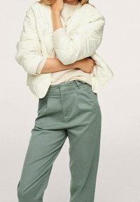Giacca bomber in crema gonfiata sopra un maglione beige chiaro, abbinata a pantaloni larghi di colore verde salvia. Le morbide trame e la vestibilità rilassata sono evidenti.