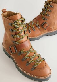 Braune Leder- und Wildleder-Wanderschuhe mit grünen und gelben Schnürsenkeln, metallischen Beschlägen und Gummisohlen. Mit einem Schnallen-Detail an der Seite.