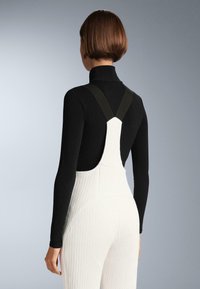 Fekete hosszú ujjú felső magas nyakrésszel, fehér, texturált kantáros nadrág alatt. A kantáros nadrág széles, fekete állítható pántokkal és gyémántmintával rendelkezik.