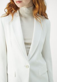 Blazer bianco su misura con un solo bottone, rever a scalpello e tasche laterali sopra un maglione a collo alto beige aderente. Tessuto dalla texture liscia.