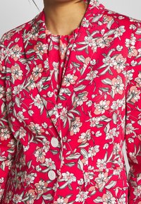 Blazer floreale con uno sfondo rosa vivace, caratterizzato da fiori bianchi e pesca, foglie verdi e due bottoni bianchi sul davanti.