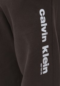 Sötétbarna pulóver fehér "Calvin Klein" felirattal és "New York City" logóval a bal ujjon. Puha, texturált anyag.