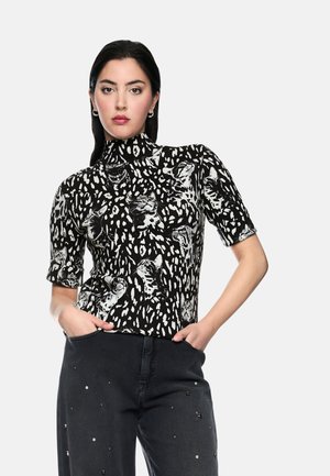 Woman with hands in pockets wearing black and white high-neck top with cat faces and spotted pattern, paired with black pants with small embellishments.