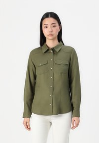 Camicia button-up verde oliva con maniche lunghe, due tasche sul petto e una texture liscia. Presenta bottoni bianchi e un colletto classico.