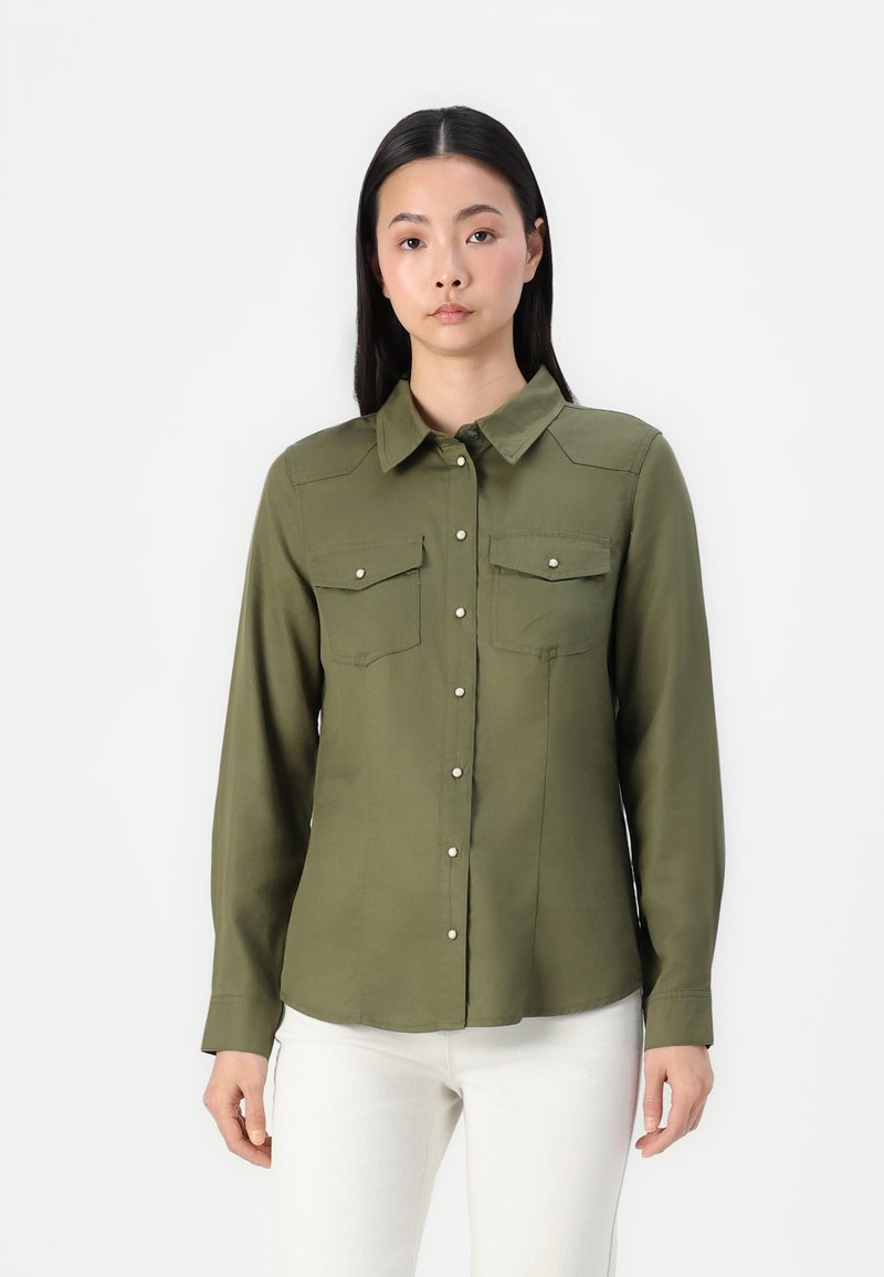 Camicia button-up verde oliva con maniche lunghe, due tasche sul petto e una texture liscia. Presenta bottoni bianchi e un colletto classico.