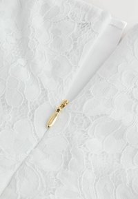 Valge pitskangas, millel on lillemustrid, ja nähtav kuldne tõmblukk. Tekstuur on õhuline ja delikaatne, rõhutades keerukaid detaile.