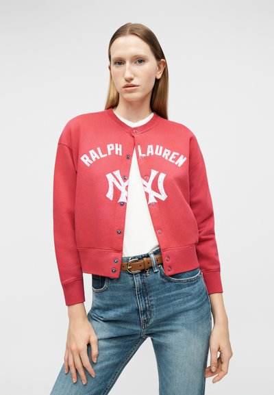Νεαρή γυναίκα που φοράει κόκκινο φούτερ Ralph Lauren με κουμπιά πάνω από λευκό πουκάμισο, μπλε τζιν με καφέ ζώνη, πόζα μπροστά σε γκρι φόντο.