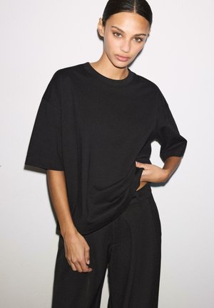 Frau mit glatt zurückgebundenem Haar, die ein übergroßes schwarzes T-Shirt und schwarze Hosen trägt, posiert vor einem schlichten weißen Hintergrund.