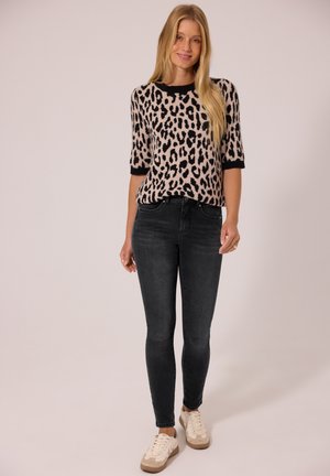 Femme aux longs cheveux blonds portant un pull à motif léopard, un jean skinny noir et des baskets blanches, debout devant un fond uni.