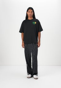 Svart oversized t-skjorte med grønn grafisk logo, sammen med svarte leopardmønstrede bukser. Hvite sneakers med grå detaljer.