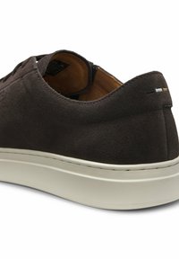 Zapatilla deportiva de ante marrón oscuro con suela de goma blanca, diseño de corte bajo, costuras sutiles en el talón en negro, blanco y beige, vista desde la parte trasera.