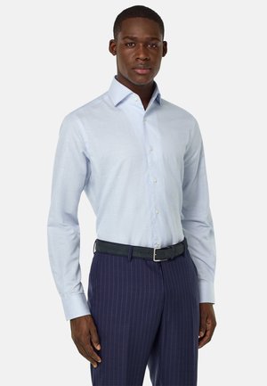 Camisa de manga larga azul claro con cuello abotonado, combinada con pantalones oscuros a rayas y un cinturón negro. Tejido suave y diseño ajustado.