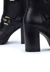 Botas de tacón de cuero negro con un tacón grueso, que cuentan con una correa de hebilla de metal dorado y detalles de costura sutiles. La suela tiene un agarre texturizado.