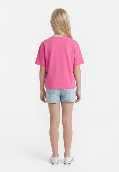 Gyerek hosszú szőke hajjal, élénk rózsaszín rövid ujjú pólóban, világoskék farmer rövidnadrágban és fehér sportcipőben, hátulnézetben.